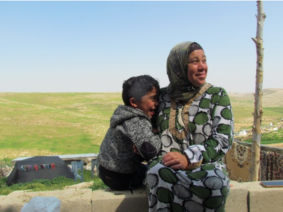 شفاء أبو عرام وابنها يجلسان في تجمُّع حلاوة، مَسافر يطا (منطقة إطلاق النار 918)  © - تصوير مكتب الأمم المتحدة لتنسيق الشؤون الإنسانية