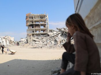 A young girl sitting on the ground with a destroyed building behind her and rubble all around. Photo by UNFPA Palestine/Media Clinic