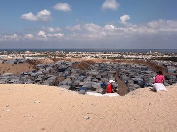 Palestinian children sit on a sandy rise overlooking a crowded displacement site in the Gaza Strip. Photo by OCHA