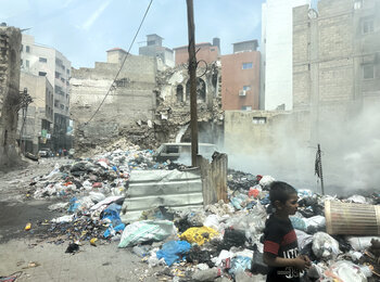 A boy walking next to scattered solid waste in a heavily damaged area of Gaza city. Photo by OCHA/Olga Cherevko, 25 April 2024