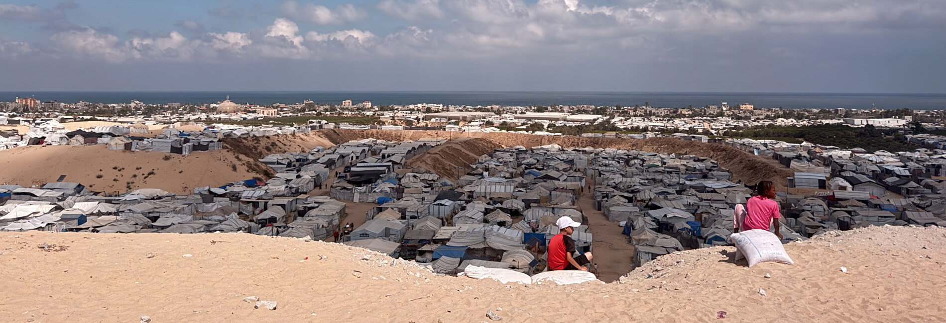 Palestinian children sit on a sandy rise overlooking a crowded displacement site in the Gaza Strip. Photo by OCHA