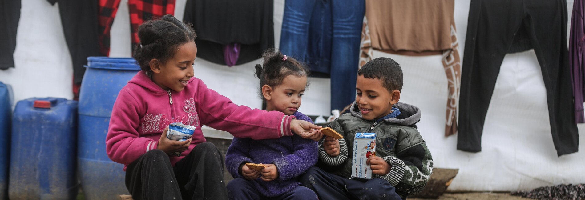 Children sitting outside their tent in Gaza, sharing biscuits delivered by the World Food Programme.  Hundreds of thousands of displaced people are now filling the streets of southern Gaza, living in makeshift shelters in miserable conditions, with little access to food, water, medicines and appropriate shelter. Photo: WFP/Ali Jadallah, January 2024 