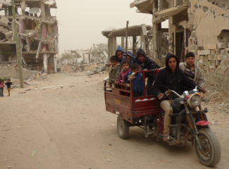 Children riding on a vehicle with damaged buildings from hostilities behind. Al Sha’af in At Tuffah area, Gaza. February 2015. Photo by OCHA